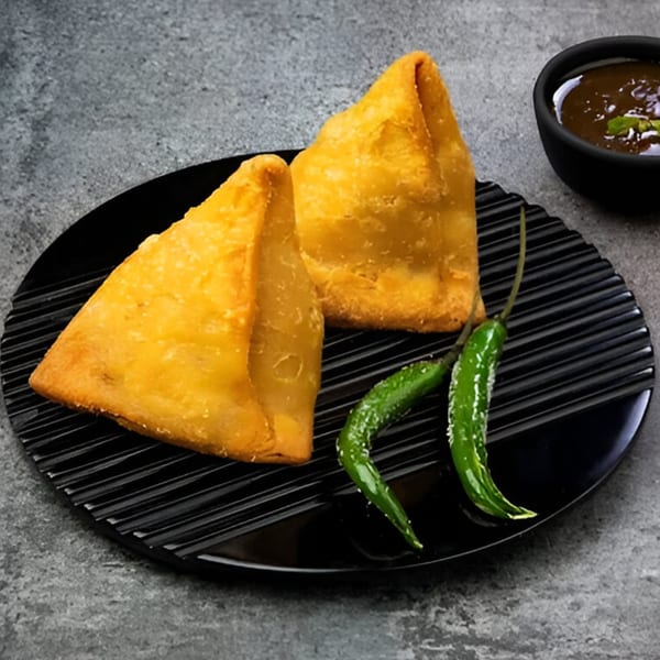Two golden-fried samosas with green chilies and a small bowl of chutney on a black plate.