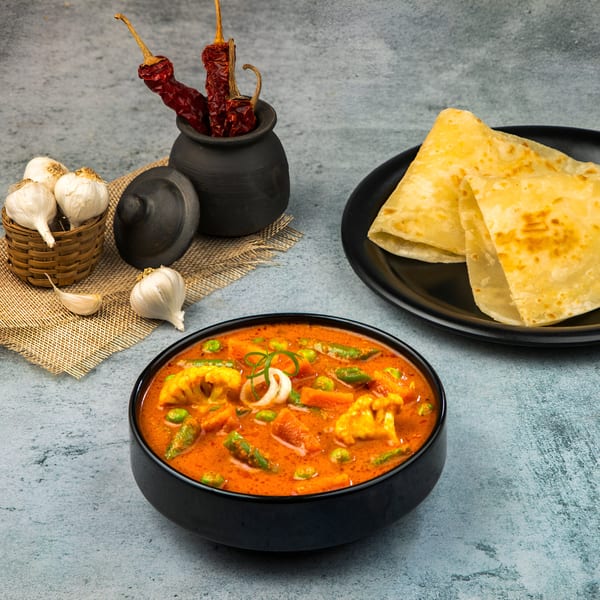 Indian vegetable curry in a black bowl with paratha bread, garlic, and dried chilies.