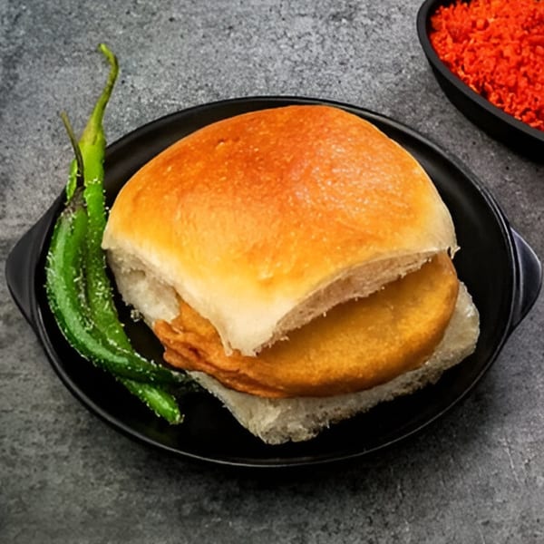A Turmerica vada pav, a fried potato patty in a bun, with green chilies and red chutney.