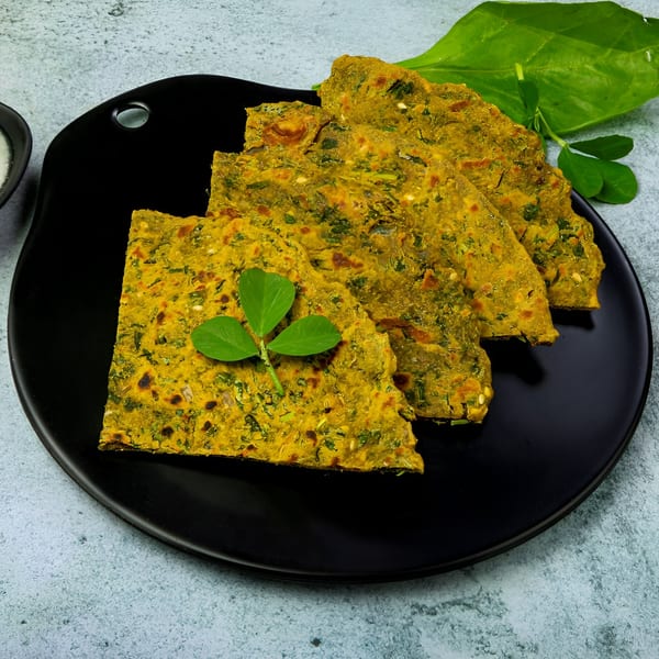 Three pieces of golden-yellow Indian flatbread, garnished with a fresh sprig of fenugreek leaves, on a black plate.